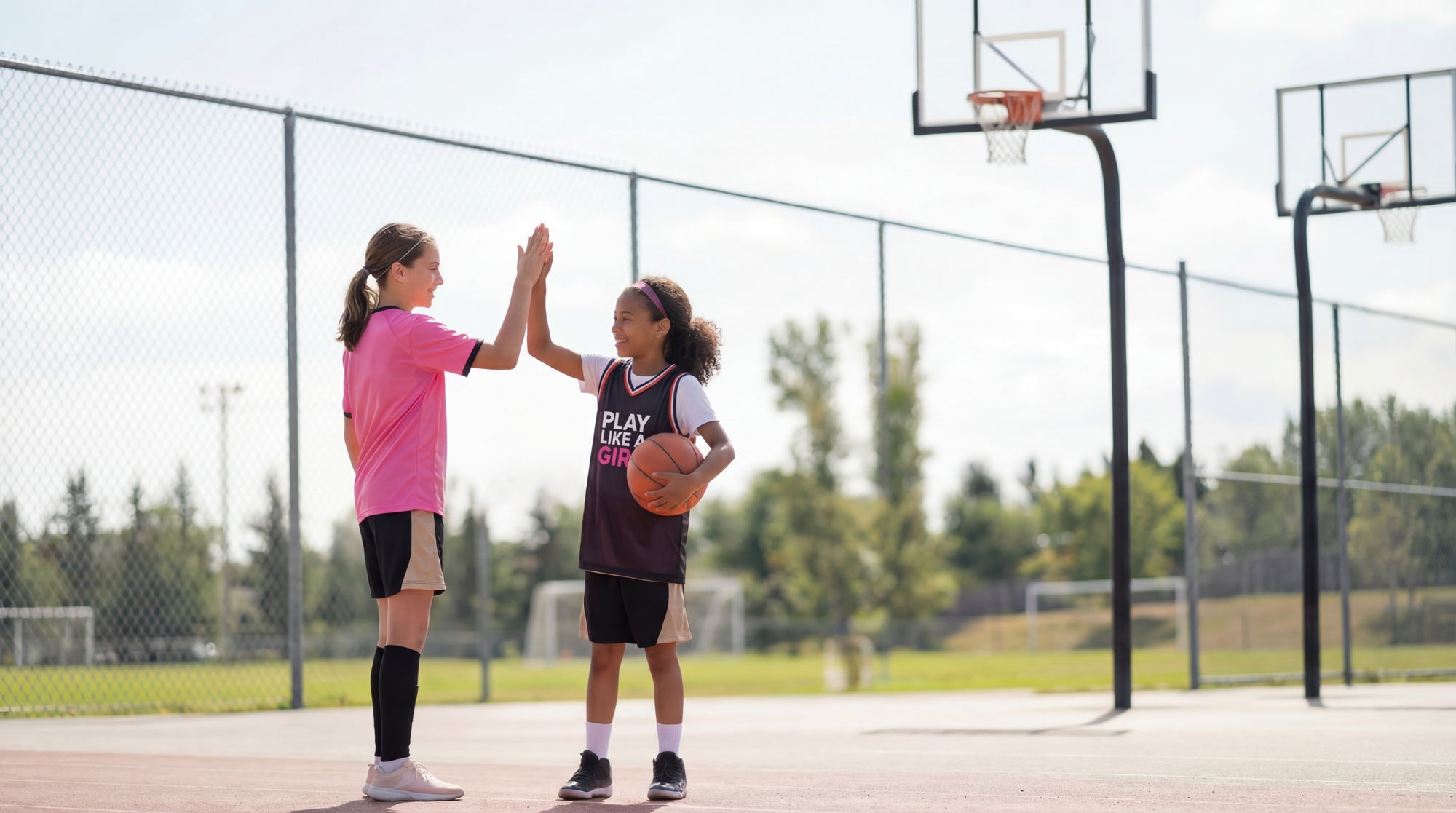 Two young girls high-fiving on a basketball court with a clear sky.